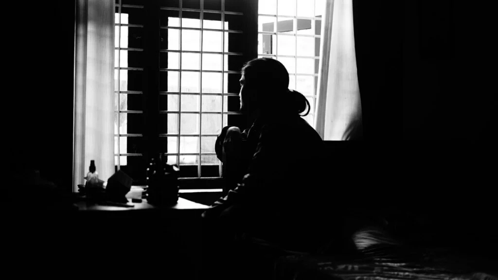 Une photographie en noir et blanc d'une personne aux cheveux longs attachés, assise dans une pièce faiblement éclairée, regardant par une fenêtre avec des barreaux métalliques. La silhouette contraste avec la lumière qui filtre à travers les rideaux. Des objets sont visibles sur une table à proximité, ajoutant à l'atmosphère contemplative.