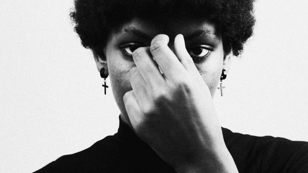 A black-and-white close-up portrait of a person with dark curly hair, looking directly at the camera. The person's right hand is raised, with the index and middle fingers touching the bridge of the nose. Black earrings with cross pendants are visible. Photograph by Jakayla Toney on Unsplash
